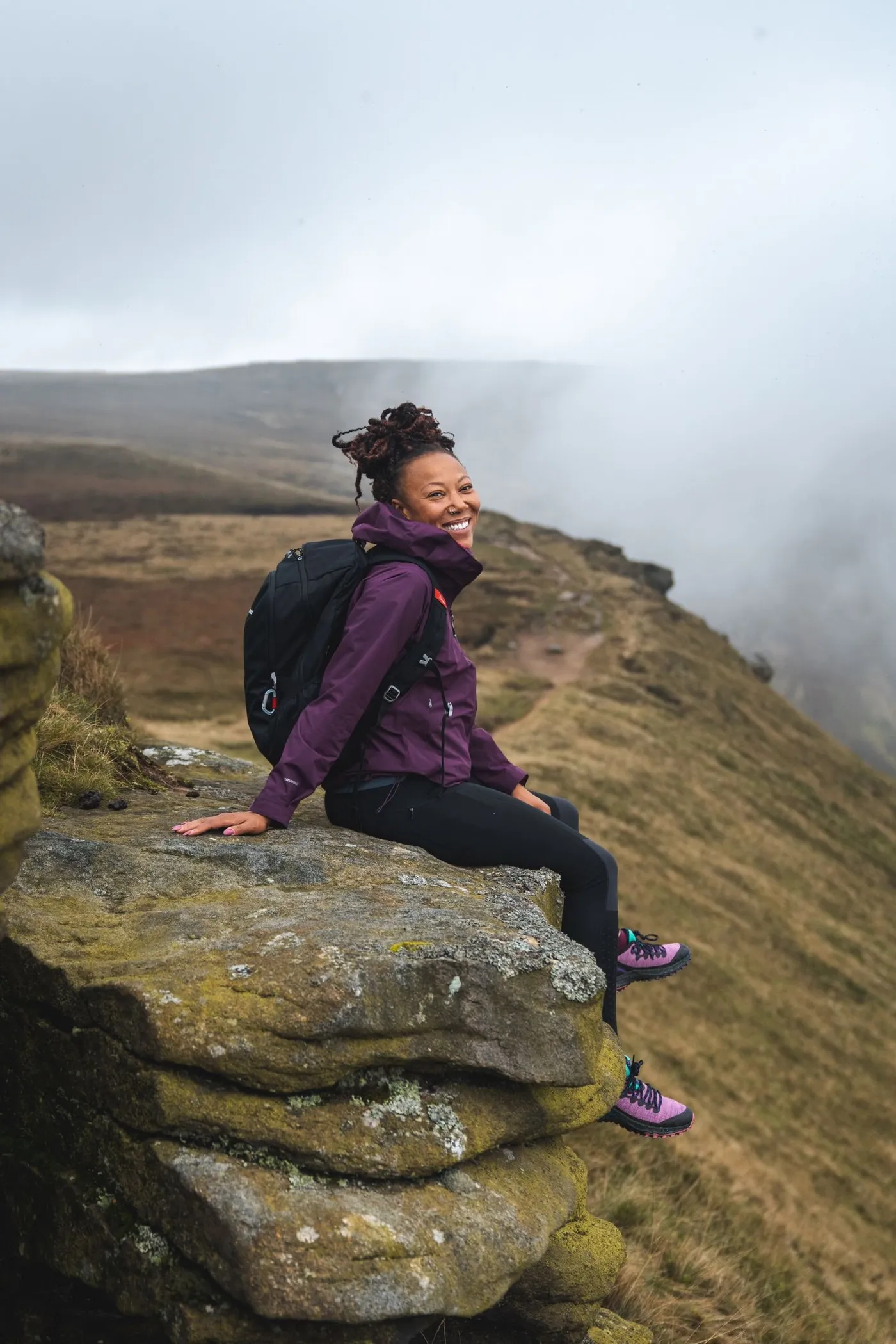 Rhiane Fatinikun sitting on a rock outcrop overlooking a misty hillside
